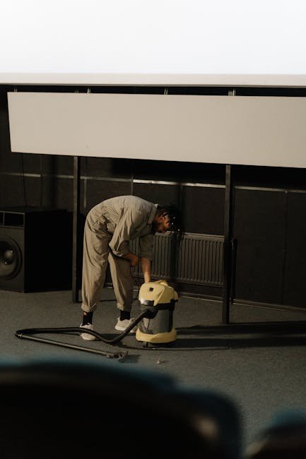 A worker in a jumpsuit using a vacuum cleaner to clean the floor of an auditorium.