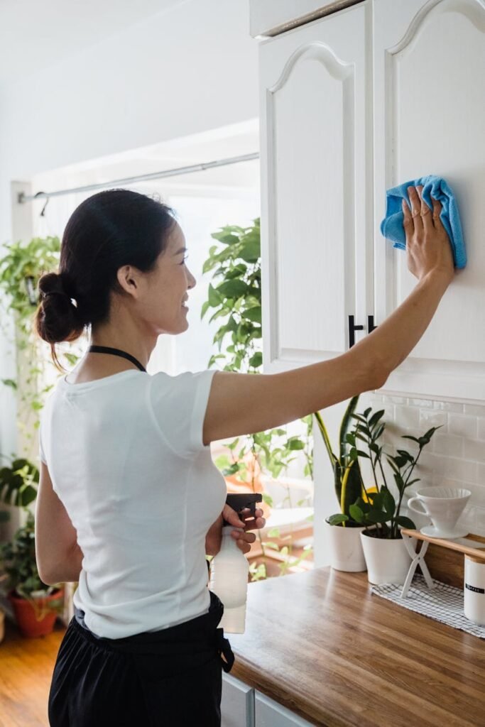 Asian woman cleaning kitchen cabinet with cloth and spray bottle indoors.